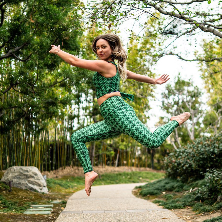 A yoga practitioner wearing green Yoga Democracy top and leggings jumps into the air.